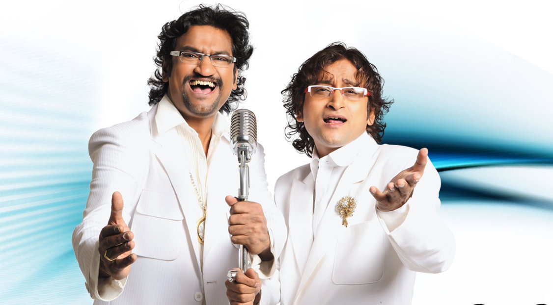 Ajay-Atul, are singing and performing in white suits with microphones, against a white and blue background.