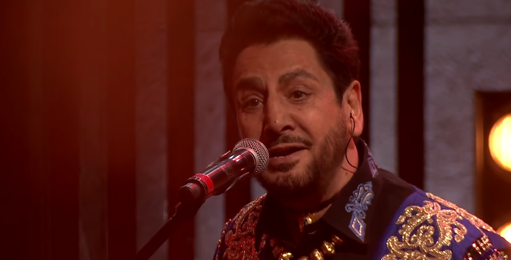 Gurdaas Maan with a mustache and goatee, wearing an ornate black shirt, singing into a microphone.