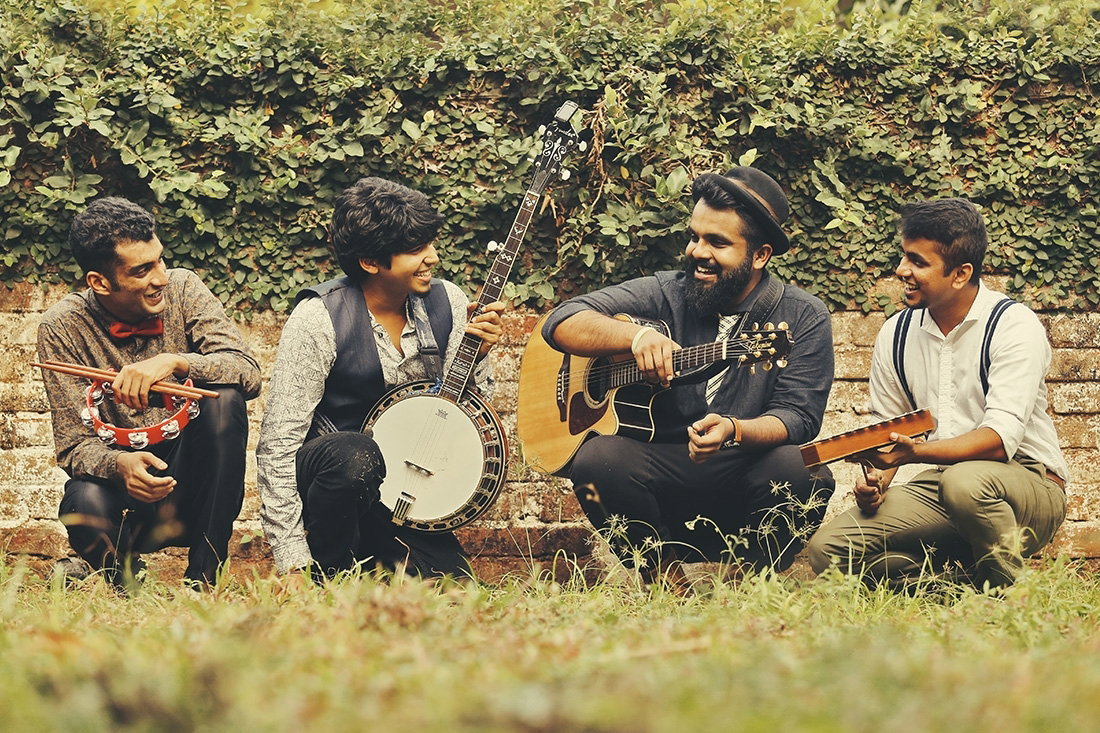When Chai Met Toast are gathered outdoors with musical instruments, smiling and looking at each other, against a leafy green background.