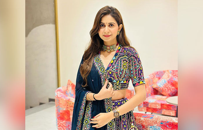  Kinjal Dave, is standing and posing in a colorful, embroidered traditional Indian outfit, looking directly at the camera.