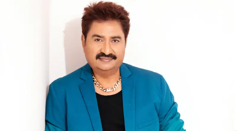 Kumar Sanu, is pictured from the chest up, smiling at the camera while wearing a blue jacket and a gold chain.