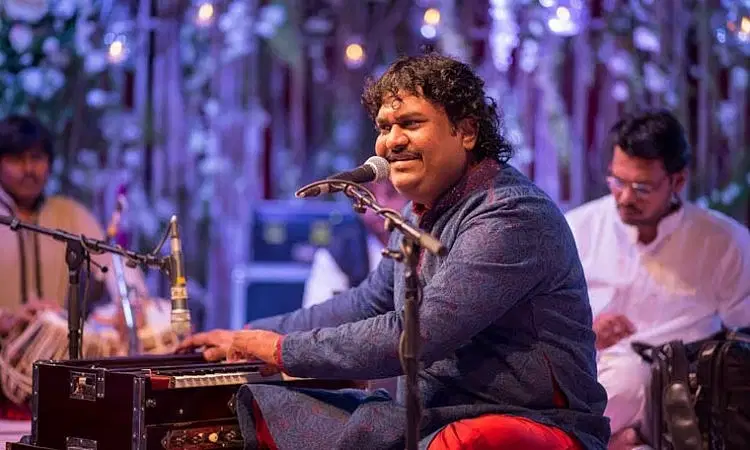 Osman Mir, is seated and performing on a harmonium with a microphone, wearing traditional attire, with other musicians in the blurred background.