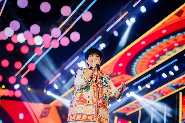 Falguni Pathak in a colorful embroidered jacket sings on a brightly lit stage with a vibrant, patterned backdrop.