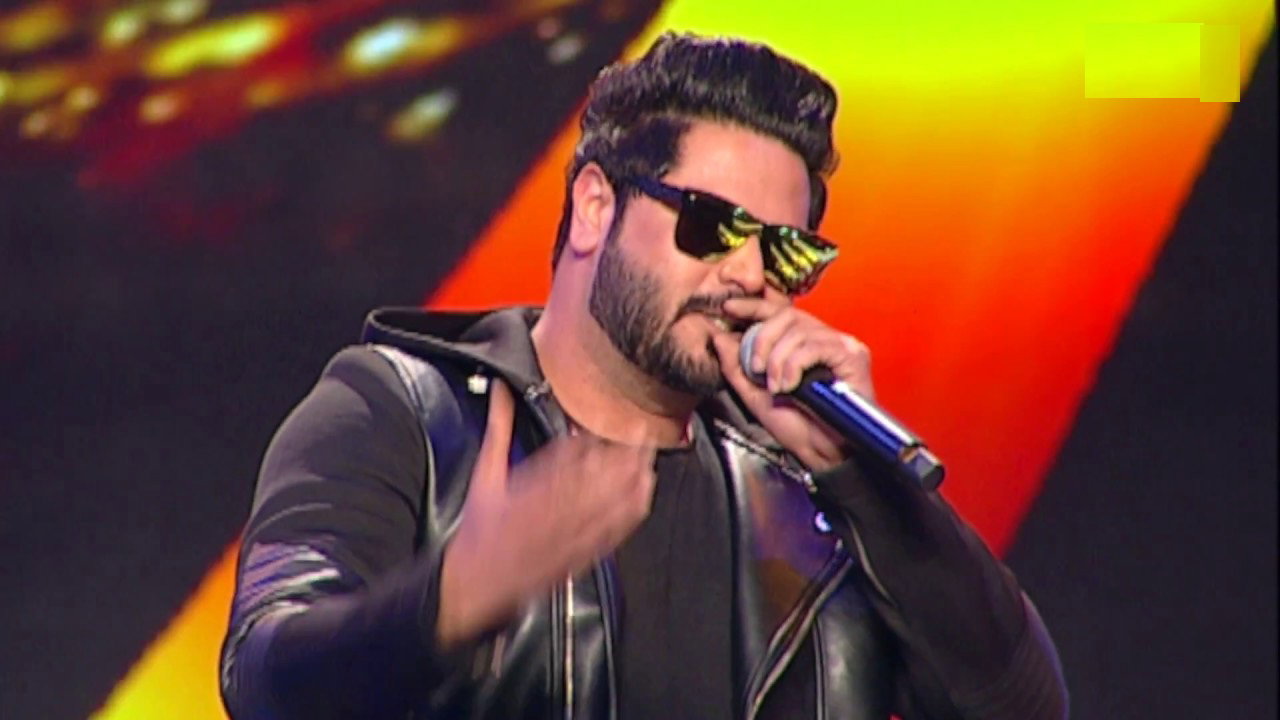 Alfaaz performs on a brightly lit stage, singing into a microphone while wearing a black jacket and reflective sunglasses.