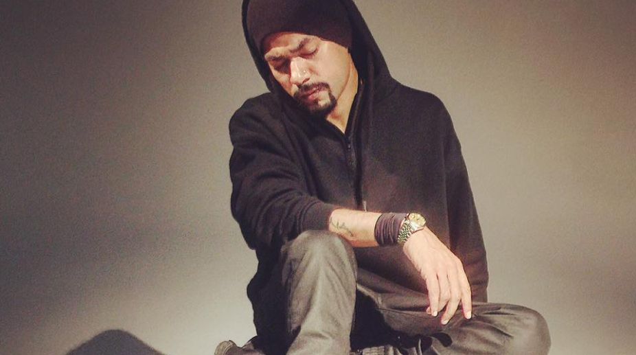 Rapper Bohemia, wearing a black hoodie with the hood up, sits in a contemplative pose with his head bowed against a plain background.