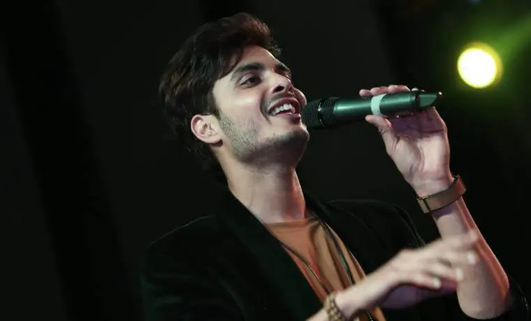 Gurnazar smiles as he sings passionately into a microphone, wearing a black blazer during a performance on a dimly lit stage.
