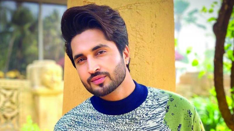 A close-up of Punjabi singer Jassi Gill with styled, voluminous hair, wearing a patterned sweater while leaning against a yellow wall.