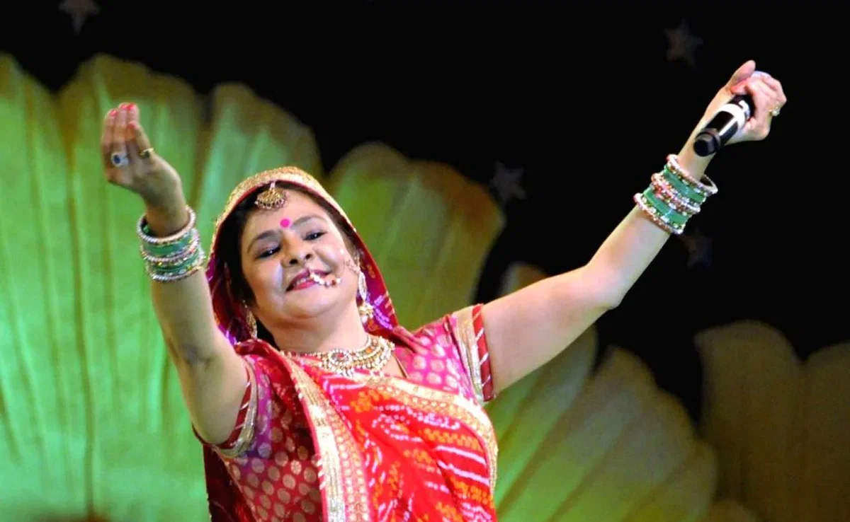 Indian folk singer Malini Awasthi, dressed in a traditional red bandhani outfit and jewelry, performs joyfully on stage with her arms raised.