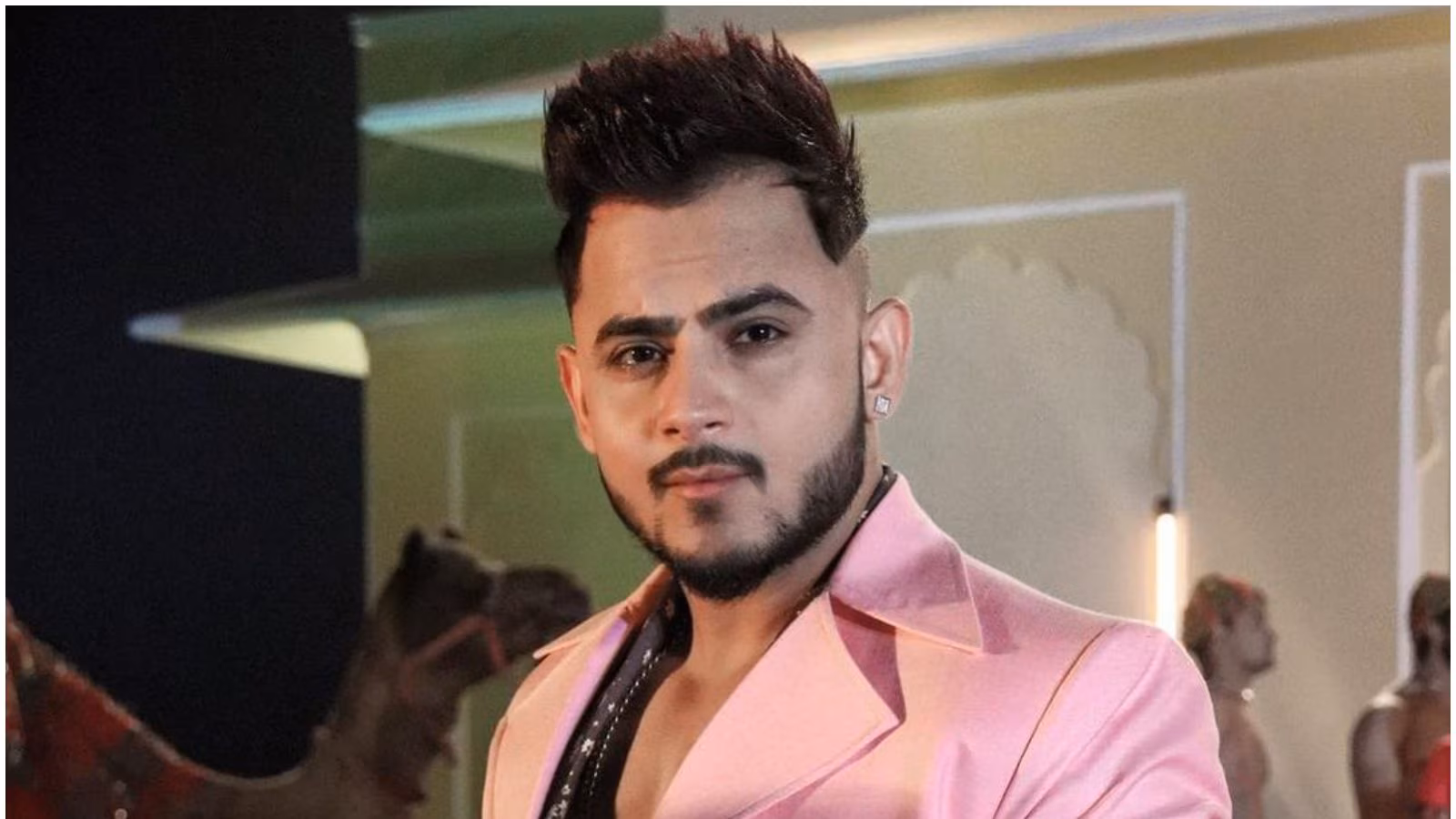 A close-up of singer Millind Gaba with a styled pompadour haircut, wearing a pink blazer over a black sequined shirt.