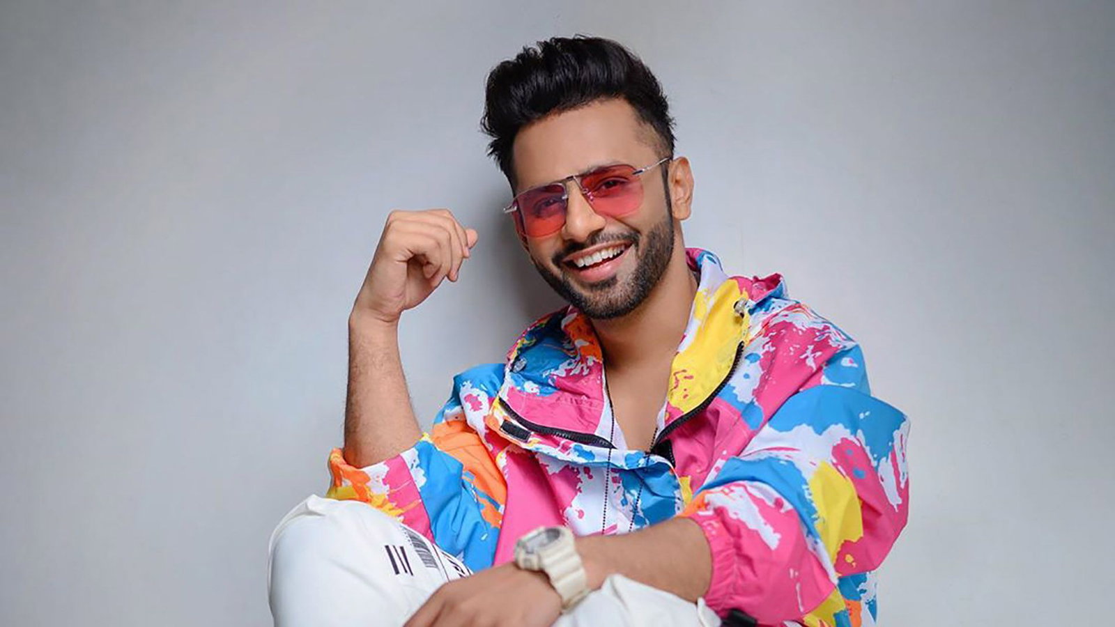 Singer Rahul Vaidya smiles at the camera, wearing a vibrant multi-colored jacket and pink-tinted sunglasses.